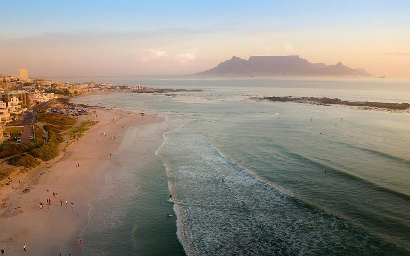 Luftaufnahme eines Sandstrandes mit Menschen, der Stadt und dem Tafelberg in der Ferne bei Sonnenuntergang.