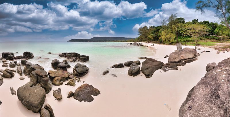 Ein breiter, heller Sandstrand mit Felsformationen im Vordergrund und im flachen Wasser, dahinter ein dichter tropischer Wald und das Meer unter einem bewölkten Himmel.