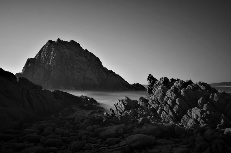 Eine felsige Küste mit einem großen Felsen im Hintergrund, umgeben von Nebel und dem Meer.