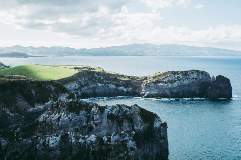 Eine felsige Kueste mit gruenen Wiesen und dem blauen Meer im Hintergrund.