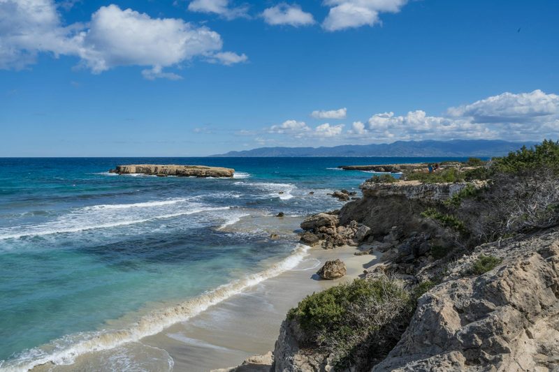 Balearen, Spanien - Eine felsige Kueste mit einem kleinen Sandstrand und tuerkisblauem Meer unter einem bewölkten Himmel.