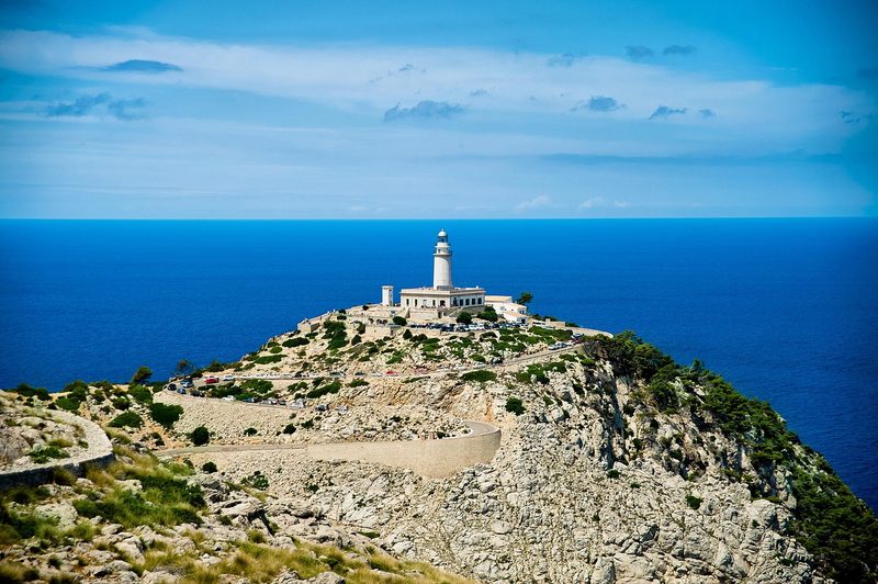 Balearen, Spanien - Ein weißer Leuchtturm steht auf einem felsigen Hügel mit Blick auf das blaue Meer.