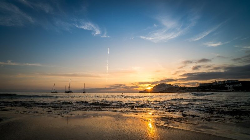 Balearen, Spanien - Ein Sonnenuntergang am Meer mit Booten im Wasser und einer Hotelanlage am Ufer.