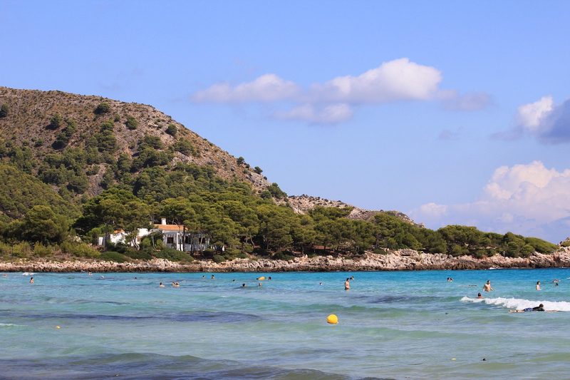 Balearen, Spanien - Menschen schwimmen und entspannen sich im türkisfarbenen Meer vor einer felsigen Küste mit Bäumen und einem weißen Haus.