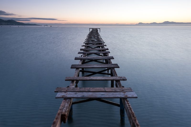 Balearen, Spanien - Ein langer, verfallener Holzsteg erstreckt sich ins ruhige Meer bei Sonnenaufgang.