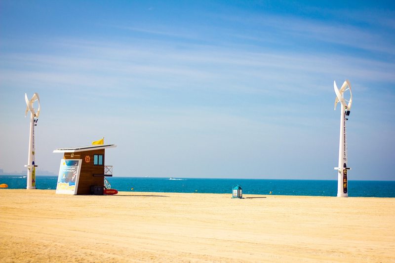 Eine Holz-Rettungsschwimmerhuette steht am Sandstrand neben einem hohen Mast mit einem Windrad und Sonnenkollektoren, im Hintergrund das blaue Meer und der Himmel.