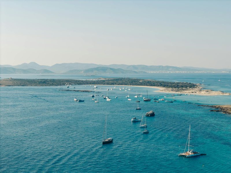 Eine Bucht mit vielen Segelbooten und Yachten vor einer Insel mit einem Sandstrand und dahinterliegenden Bergen.
