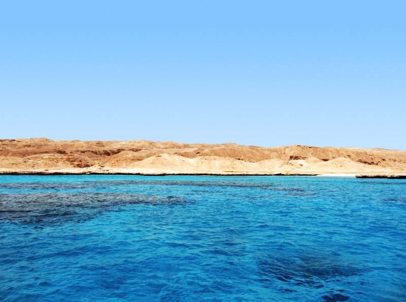 Ein Blick auf das tuerkisblaue Meer mit einer felsigen Wuestenlandschaft im Hintergrund unter einem klaren blauen Himmel.