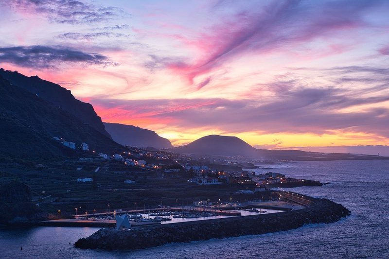 Kanaren, Spanien - Ein Hafen mit Booten bei Sonnenuntergang, mit einer Stadt, die sich an einen Berghang schmiegt, und dem Meer im Vordergrund.