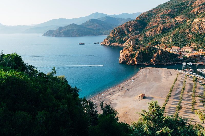 Ein Kiesstrand mit wenigen Menschen und einem kleinen Gebaeude, daneben ein Hafen mit vielen Booten, im Hintergrund eine felsige Kueste mit einem Dorf und Berge.