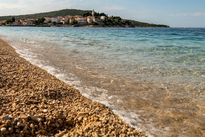 Ein Kiesstrand mit klarem, türkisfarbenem Wasser und einem Küstenort im Hintergrund.