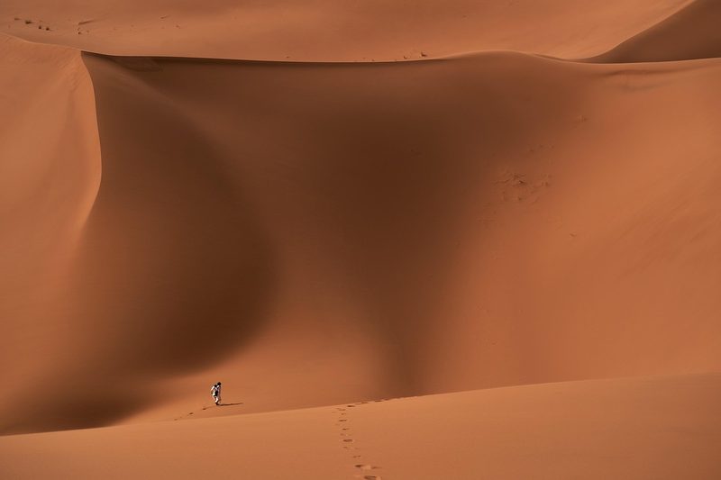 Eine Person wandert durch eine weite Dünenlandschaft in der Wüste und hinterlässt Spuren im Sand.