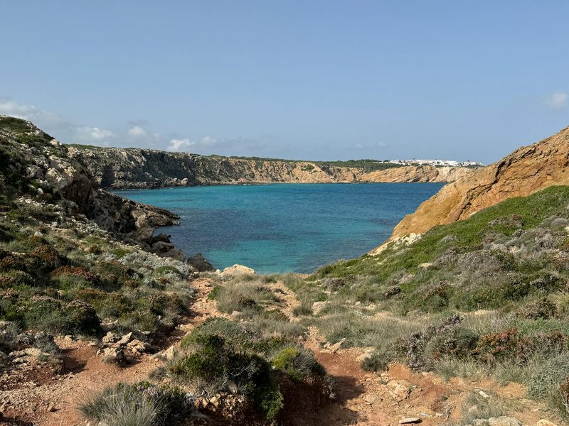 Ein Blick auf eine felsige Bucht mit tuerkisem Wasser, umgeben von mediterraner Vegetation und Felsen, mit einer kleinen Siedlung am Horizont.