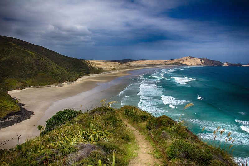 Ein langer Sandstrand mit tuerkisblauem Meer und Wellen, flankiert von gruenen Huegeln und Sandduenen.