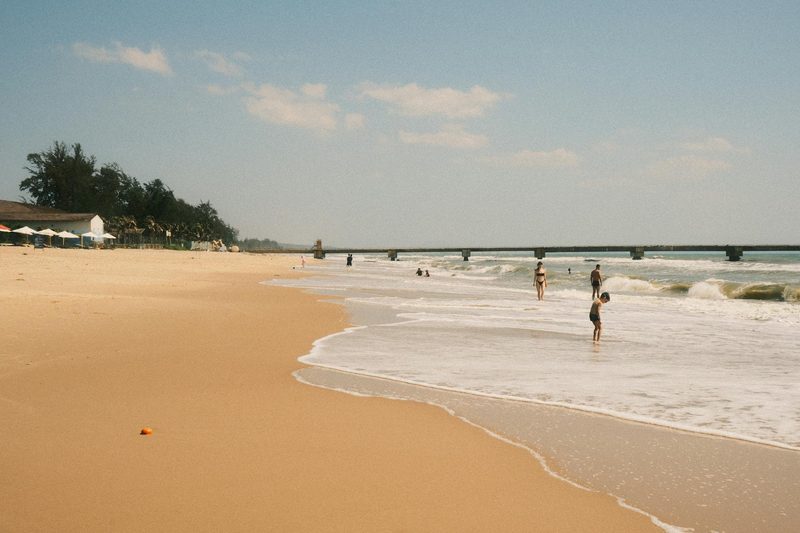 Ein breiter Sandstrand mit Menschen, die im Meer baden, und einem langen Pier im Hintergrund.