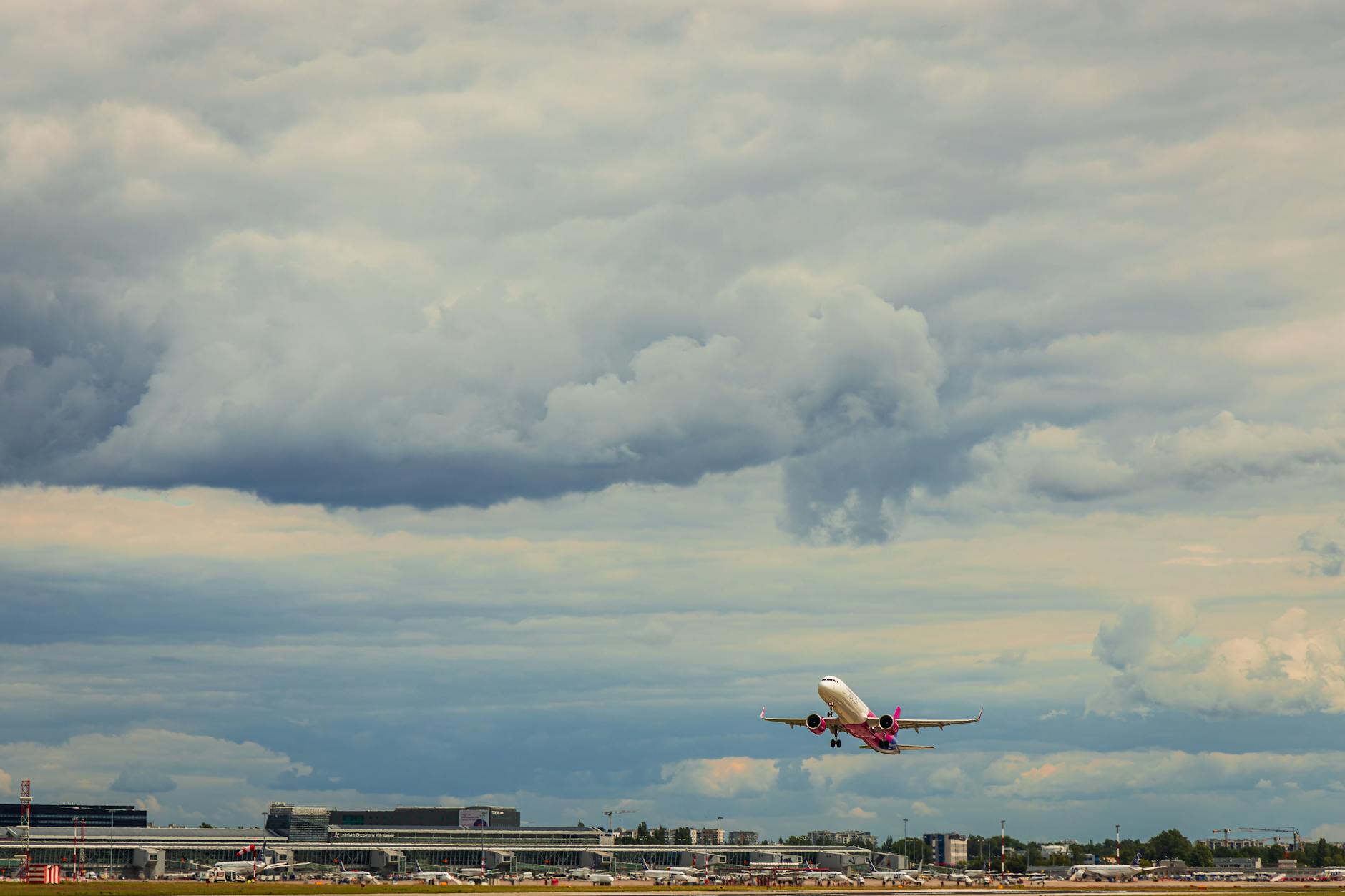 Ein weißes Flugzeug mit rosa Akzenten hebt vom Boden ab, mit einem Flughafen und bewölktem Himmel im Hintergrund.