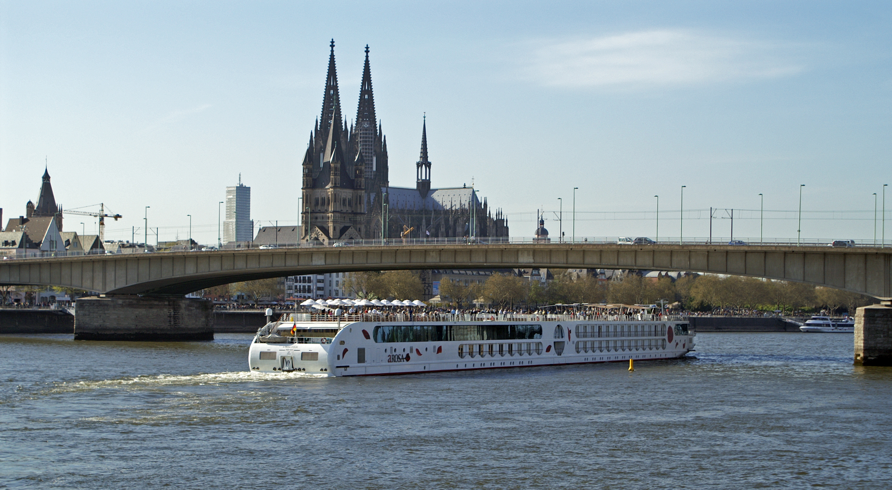 Ein Flusskreuzfahrtschiff faehrt auf einem Fluss unter einer Bruecke hindurch, im Hintergrund ist der Kölner Dom zu sehen.