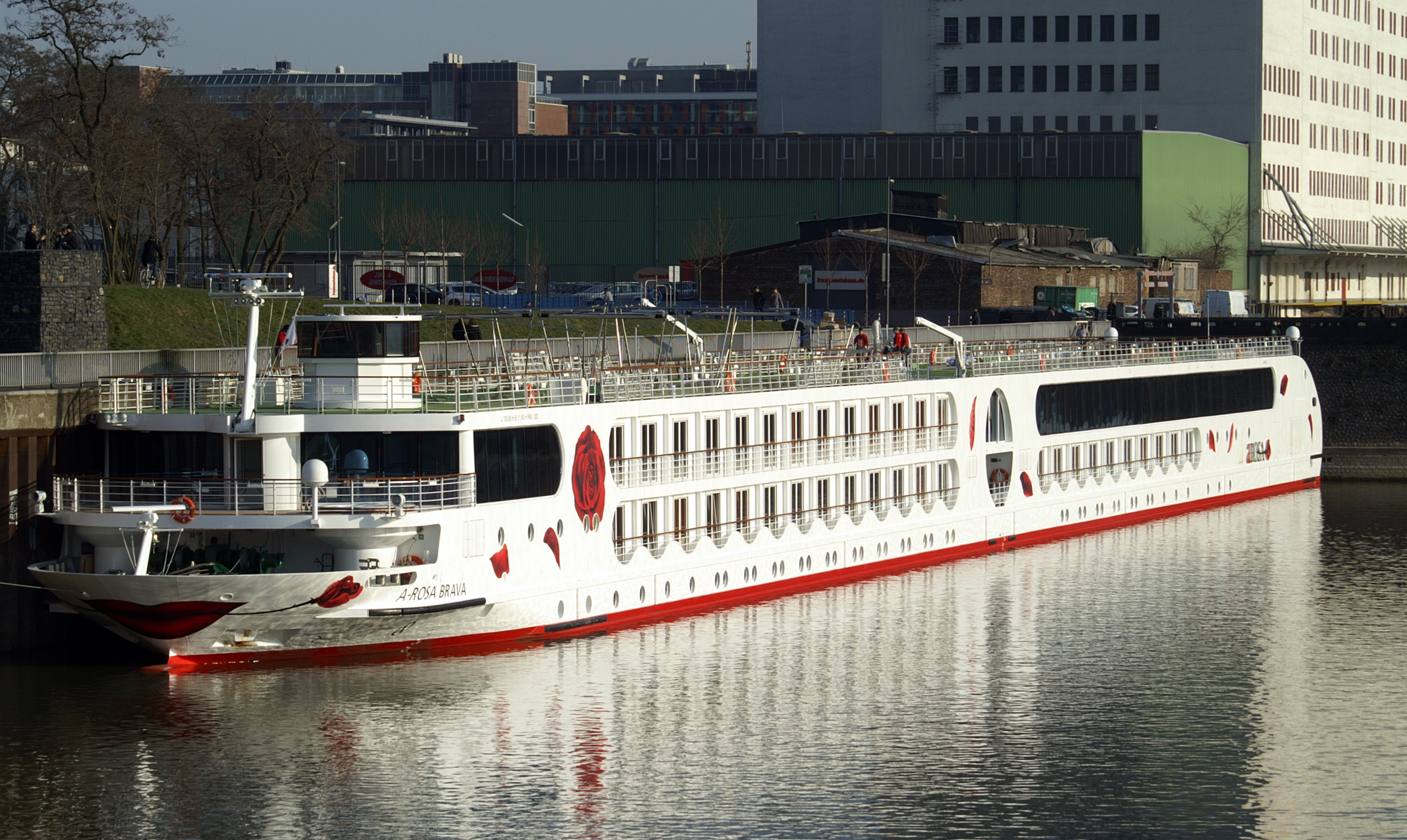 Ein weisses Flusskreuzfahrtschiff mit rotem Rumpf und roten Lippenmotiven am Bug liegt im Wasser, im Hintergrund sind Gebäude einer Stadt zu sehen.