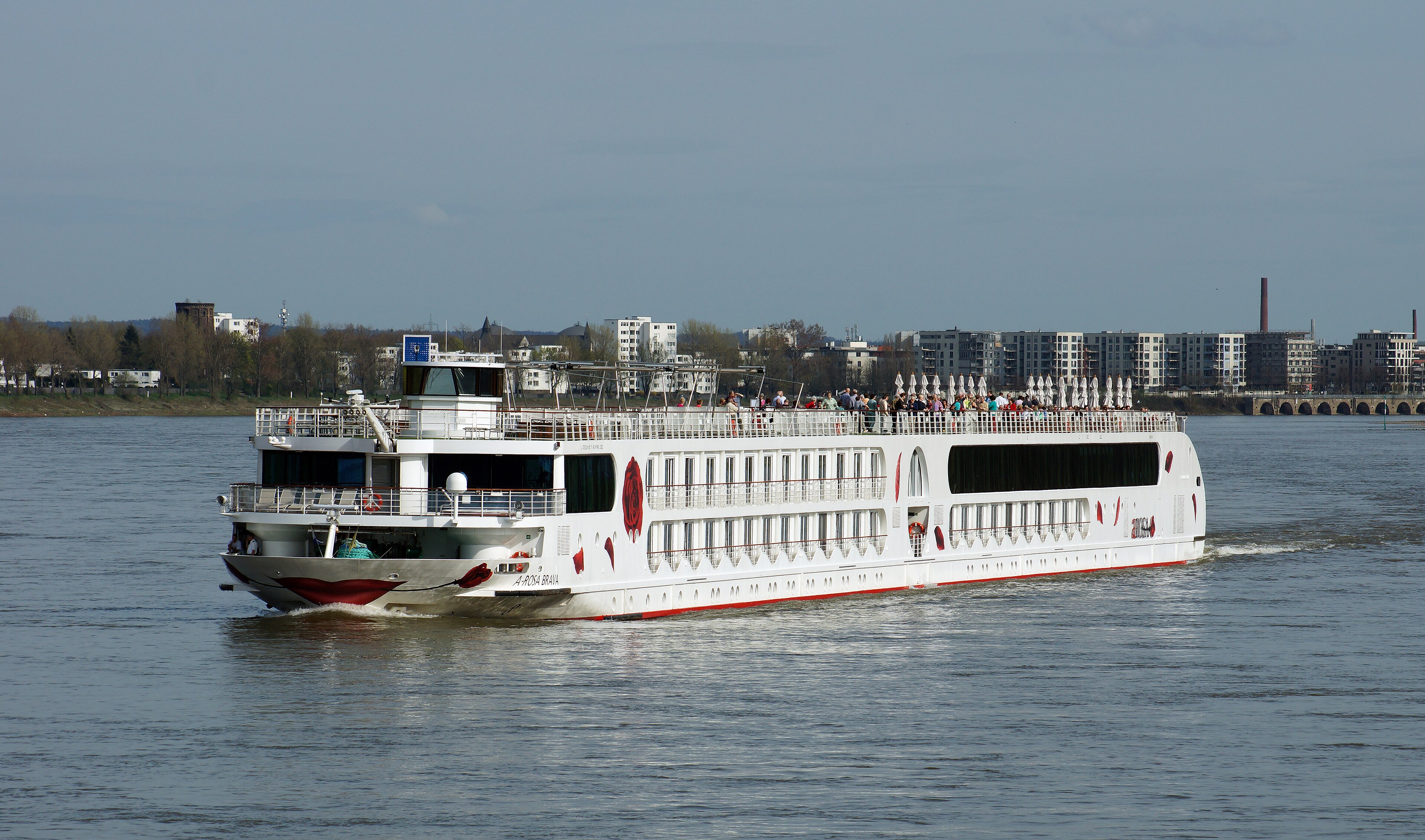 Ein weisses Flusskreuzfahrtschiff mit dem Namen A-ROSA BRAVA faehrt auf einem Fluss, im Hintergrund eine Stadtkulisse mit Gebaeuden und einer Bruecke.