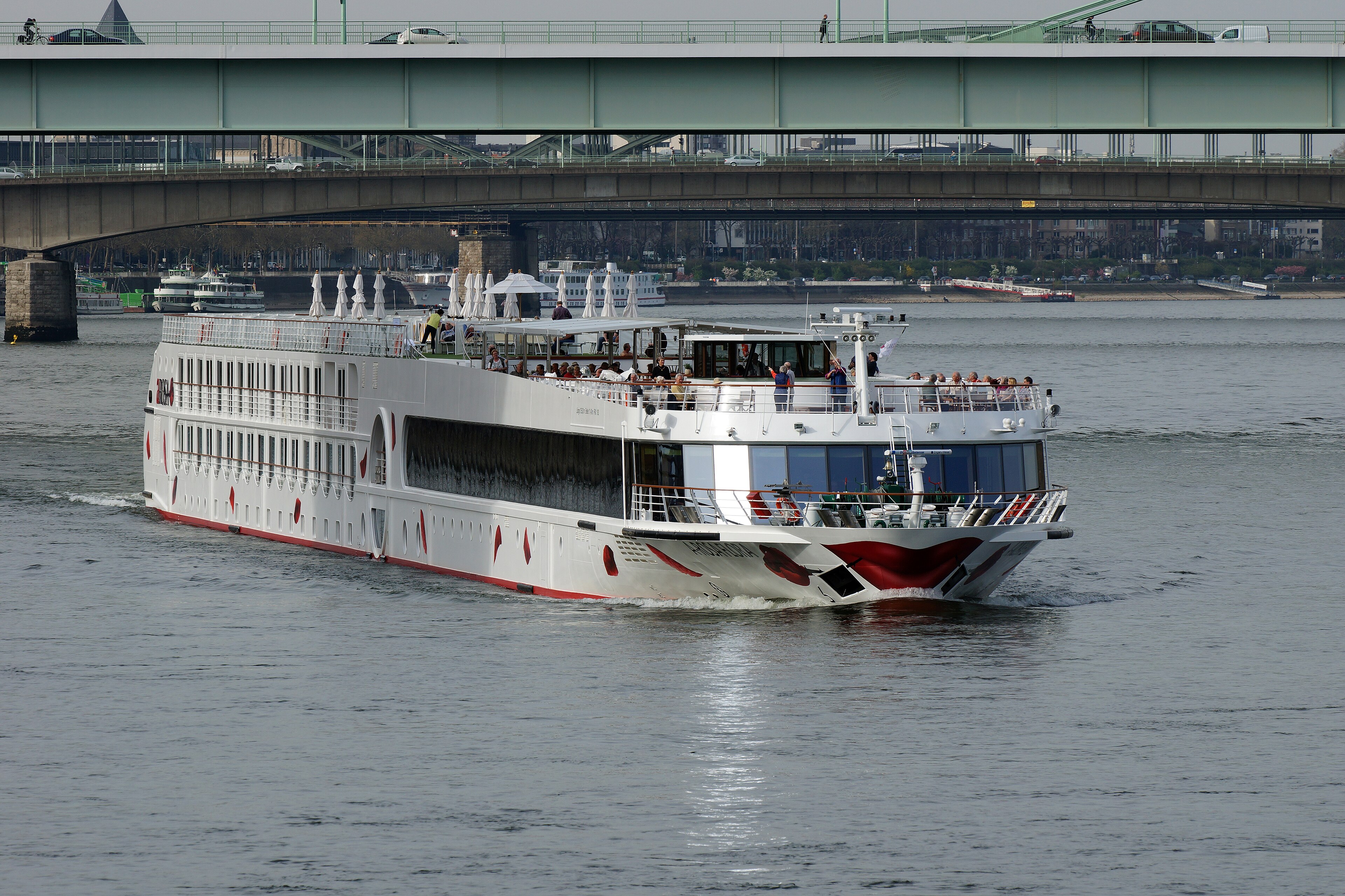 Ein weißes Flusskreuzfahrtschiff mit roten Akzenten fährt auf einem Fluss unter mehreren Brücken hindurch, auf denen Autos und Fußgänger zu sehen sind.