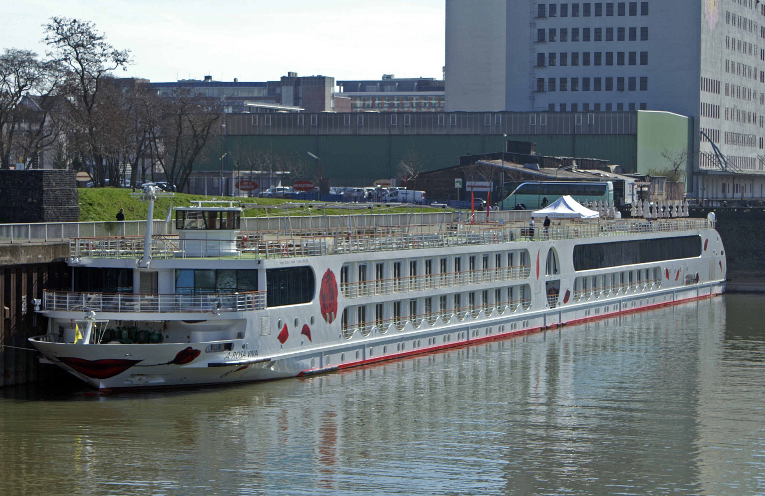 Ein weisses Flusskreuzfahrtschiff mit dem Namen A-ROSA VIVA liegt an einer Kaimauer in einer Stadt.
