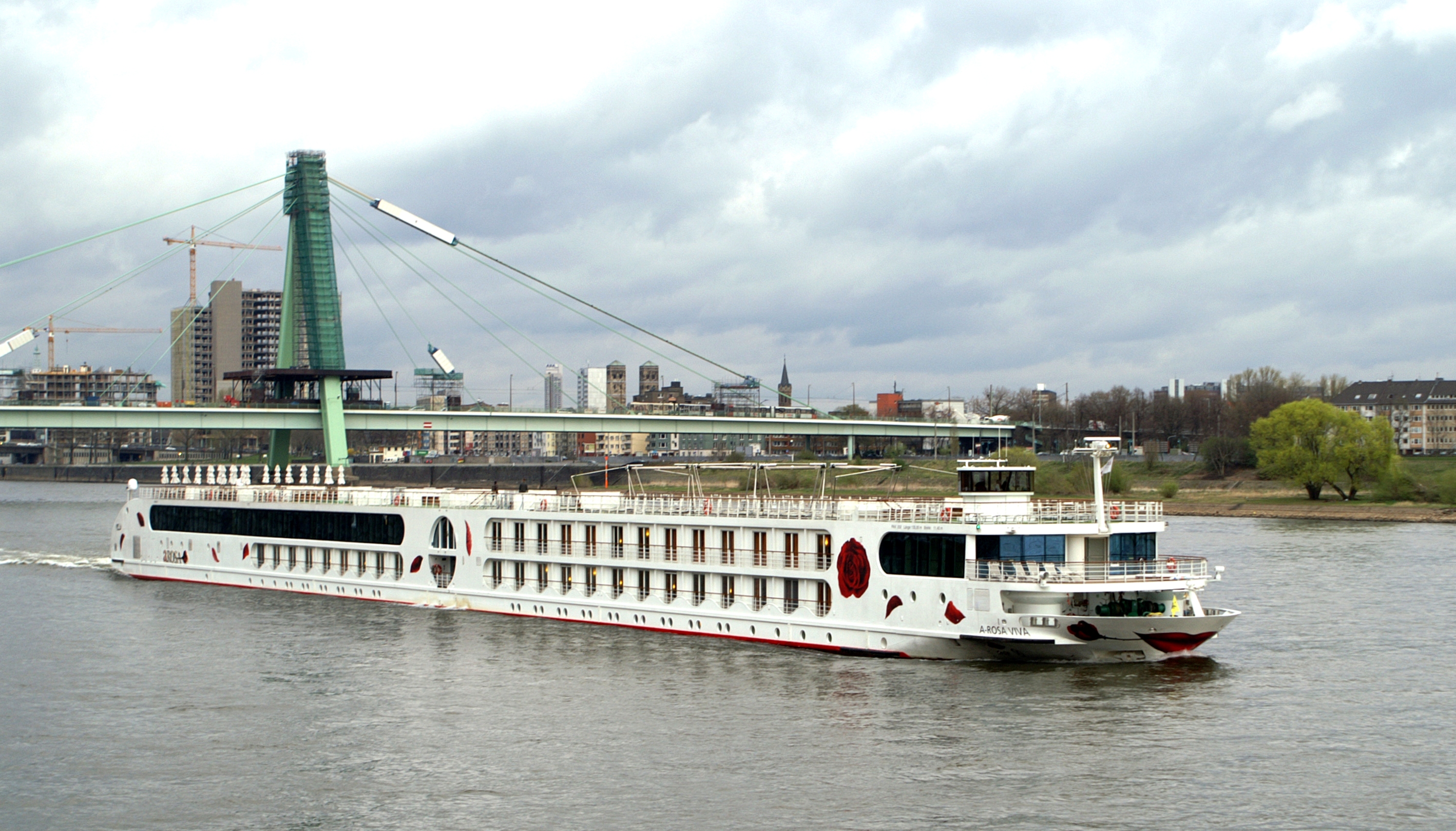 Ein weißes Flusskreuzfahrtschiff mit dem Namen A-ROSA VIVA fährt auf einem Fluss, im Hintergrund eine Stadt und eine Brücke.