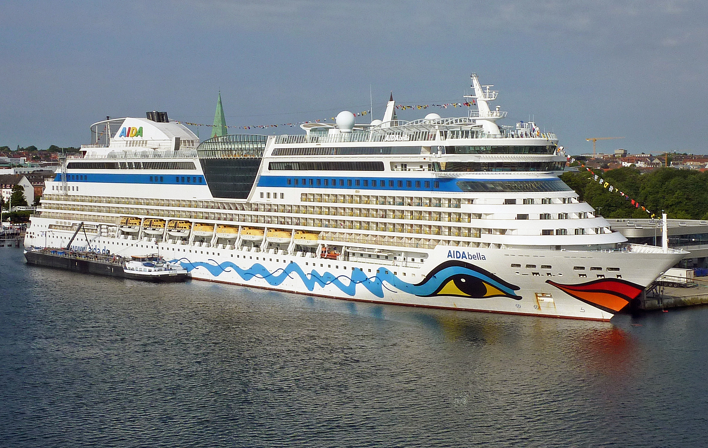 Ein großes weißes Kreuzfahrtschiff mit dem Namen AIDAbella und einem bunten Auge-Motiv am Bug liegt im Hafen.