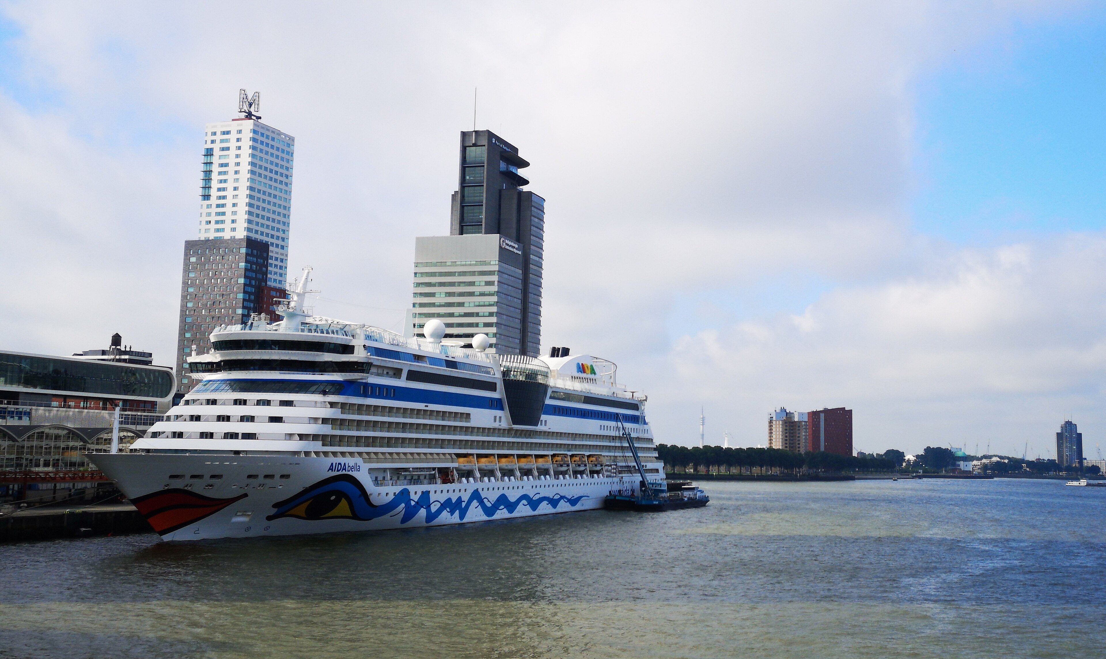 Ein großes Kreuzfahrtschiff liegt im Hafen vor einer Stadtkulisse mit mehreren Hochhäusern.
