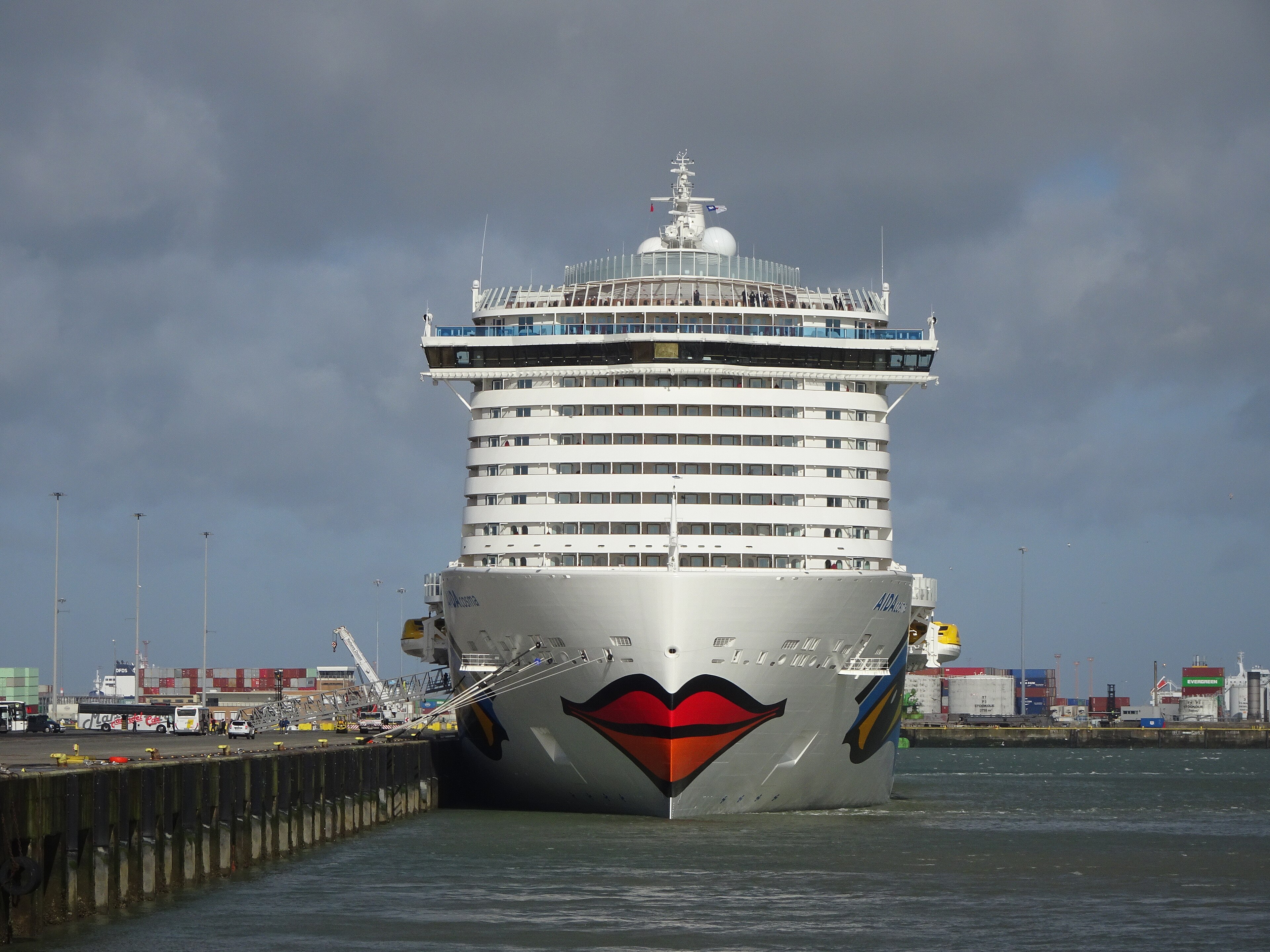 Ein großes Kreuzfahrtschiff mit dem bekannten AIDA-Kussmund-Motiv liegt im Hafen vor einer Kulisse aus Containern und Kränen.