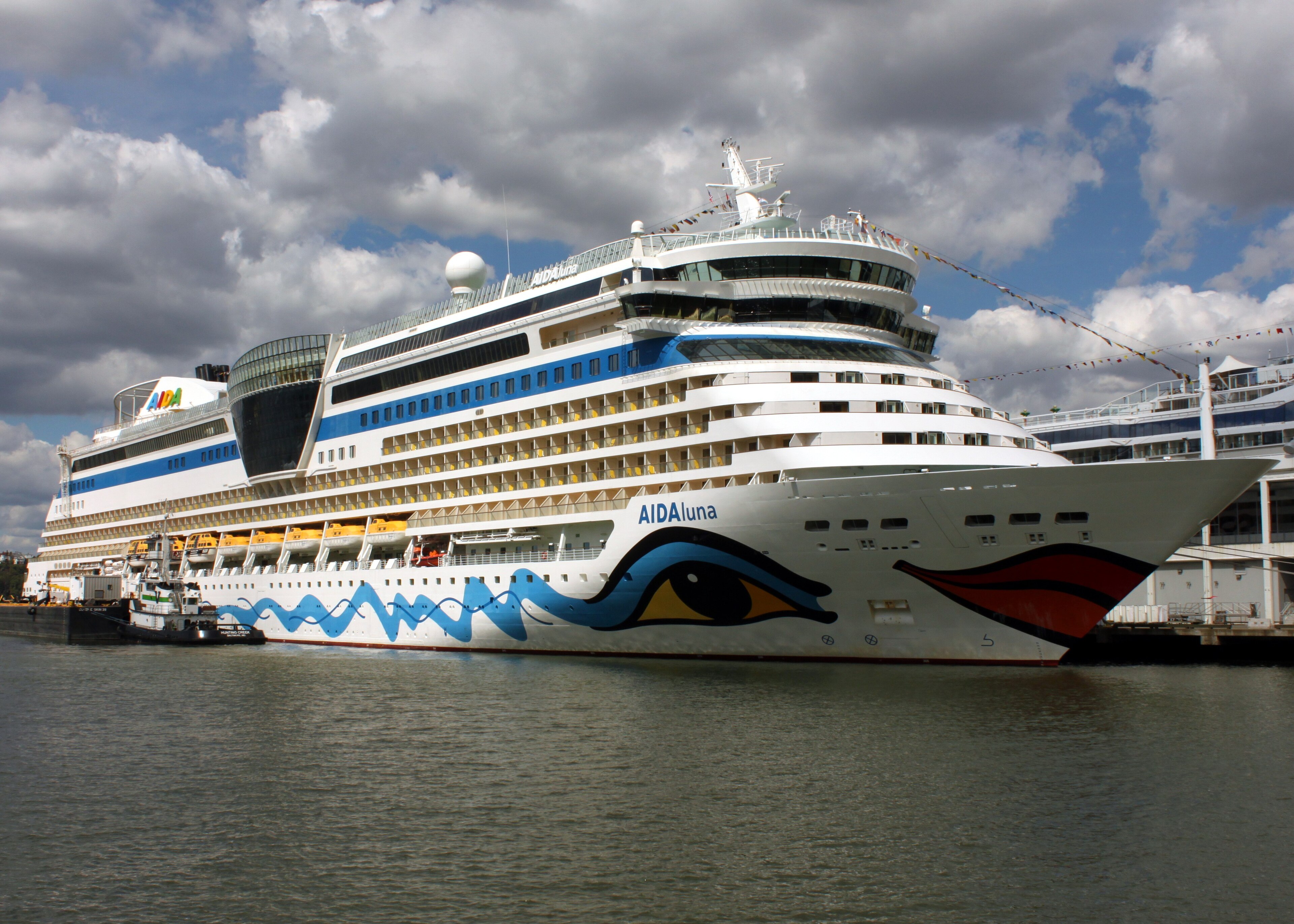 Ein großes Kreuzfahrtschiff mit dem Namen AIDAluna liegt im Hafen vor einer bewölkten blauen Himmelskulisse.