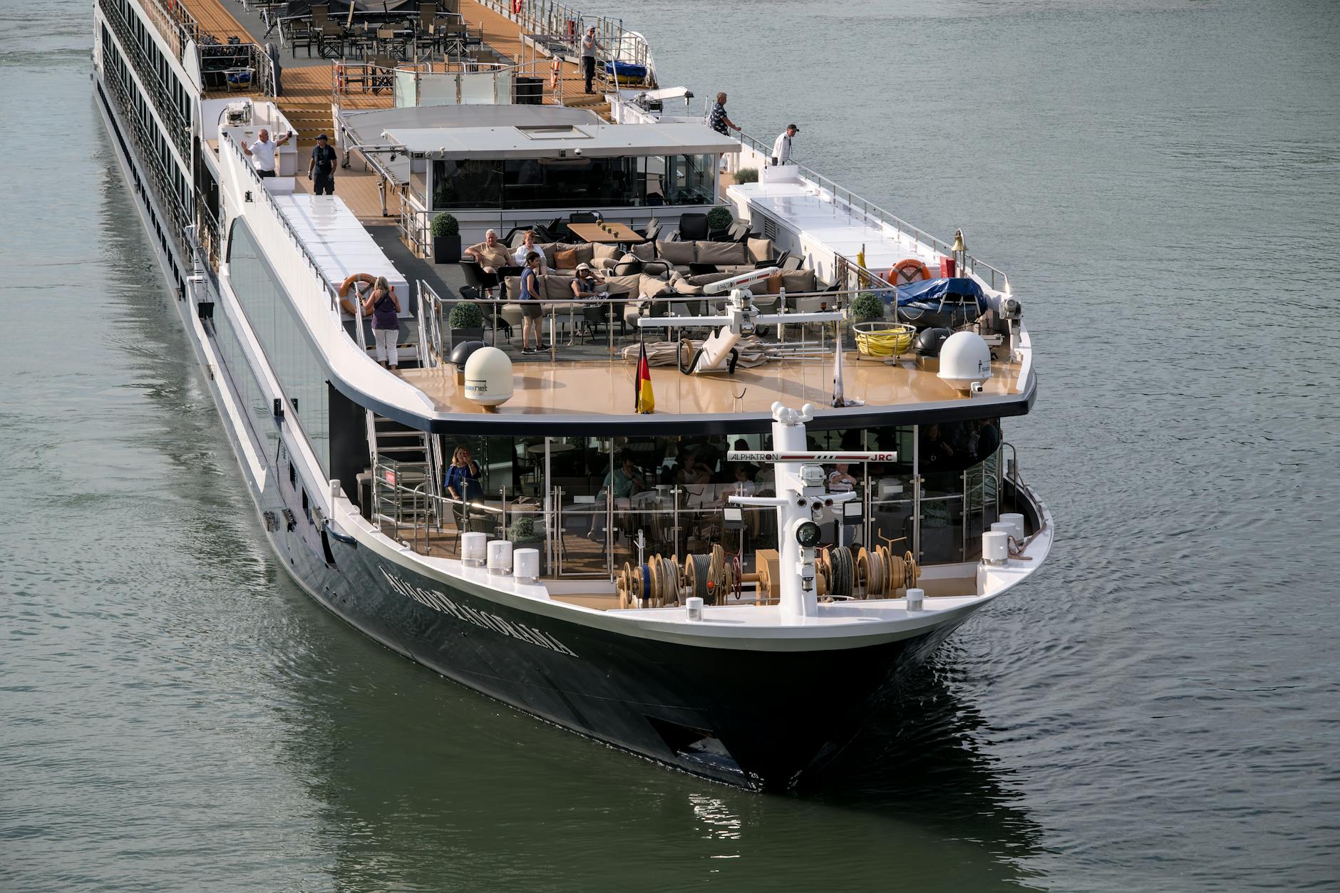 Ein Flusskreuzfahrtschiff mit Passagieren auf dem Oberdeck fährt auf einem Fluss.