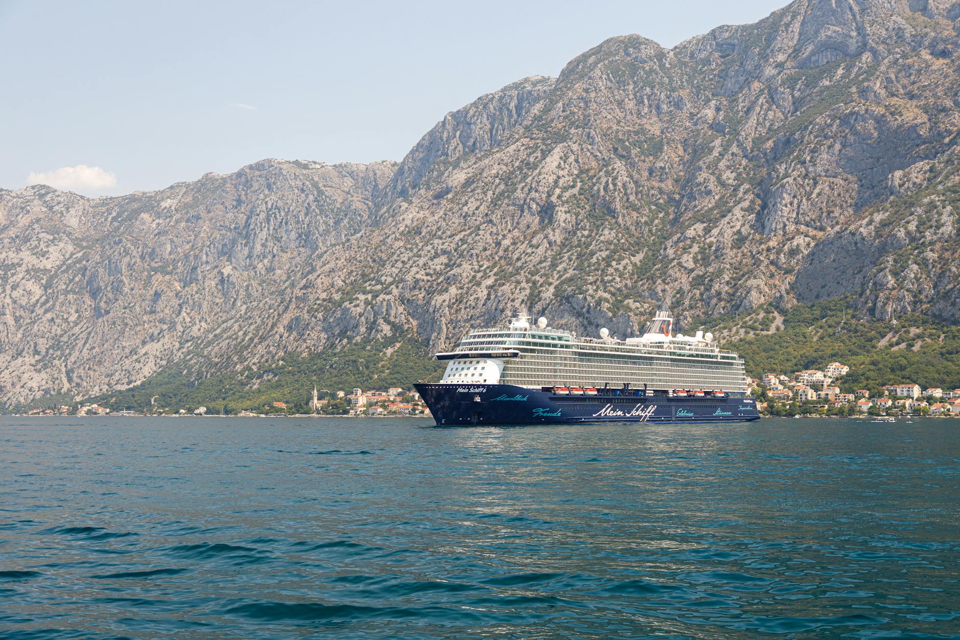 Ein großes Kreuzfahrtschiff fährt auf dem Meer vor einer Berglandschaft mit einer Küstenstadt.