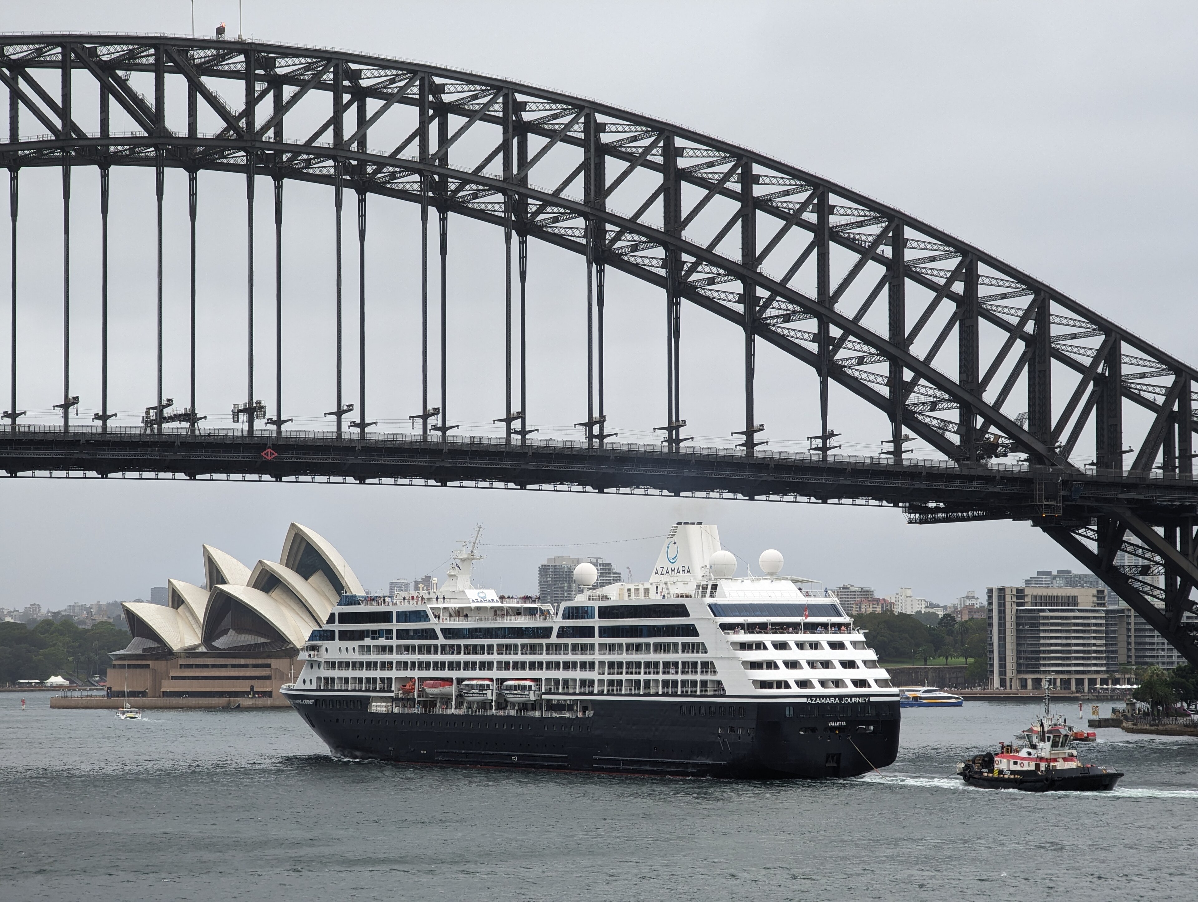 Ein Kreuzfahrtschiff faehrt unter der Sydney Harbour Bridge hindurch, im Hintergrund ist die Sydney Opera zu sehen.