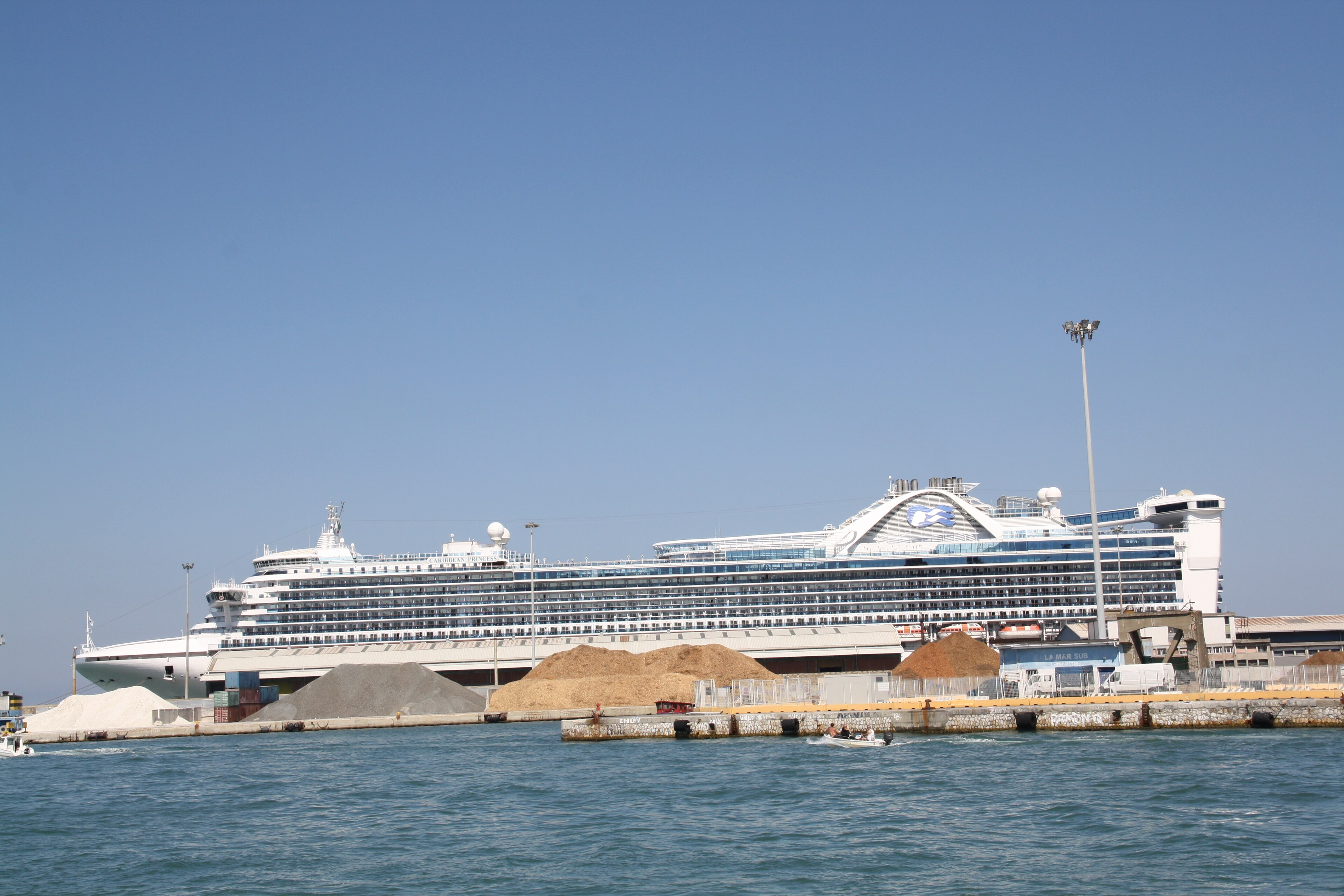Ein großes weißes Kreuzfahrtschiff liegt im Hafen vor einer Kulisse aus Gebäuden und Lagerhäusern.
