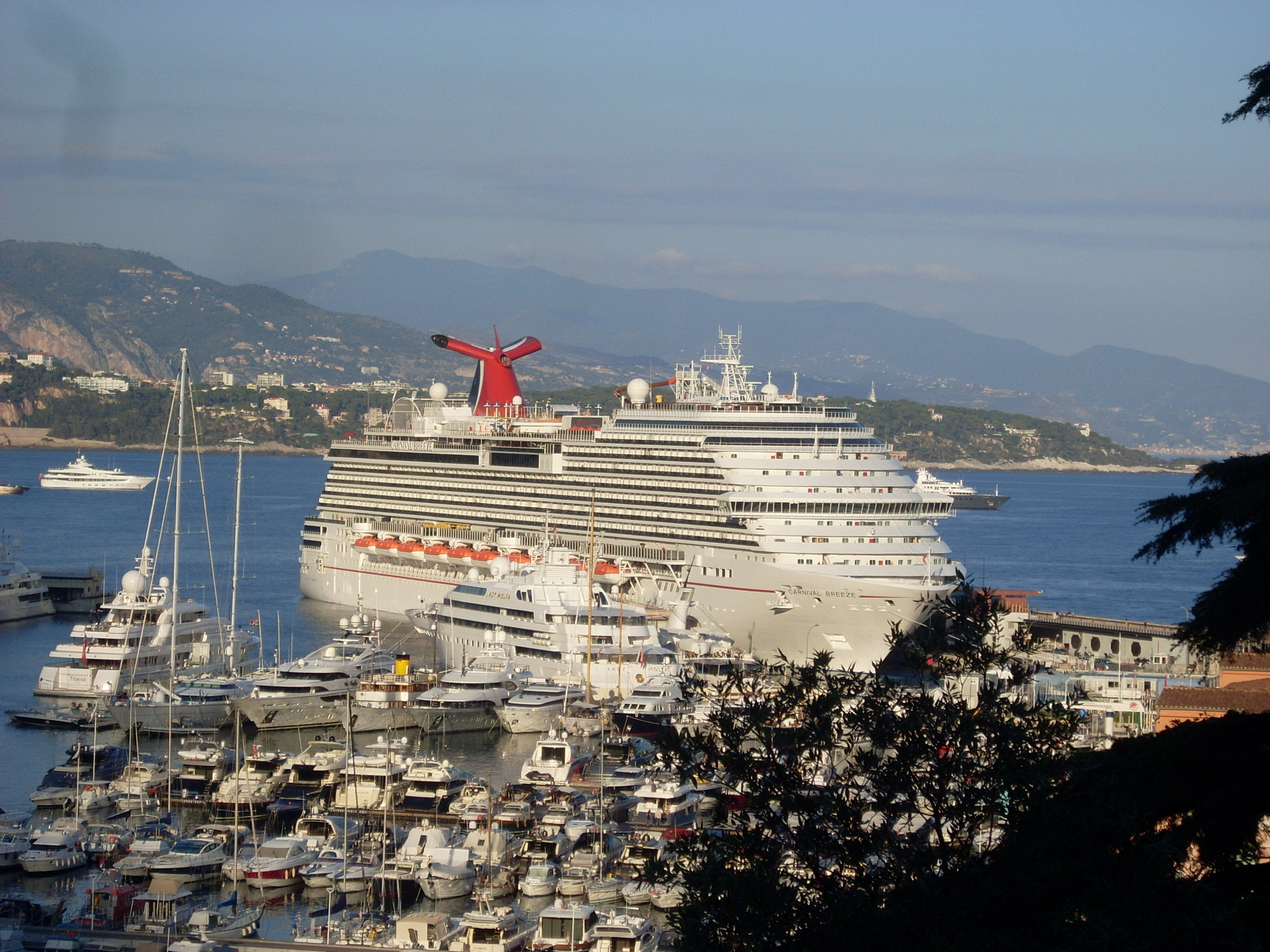 Ein grosses Kreuzfahrtschiff liegt im Hafen vor einer bergigen Kuestenlandschaft, umgeben von vielen kleineren Yachten.