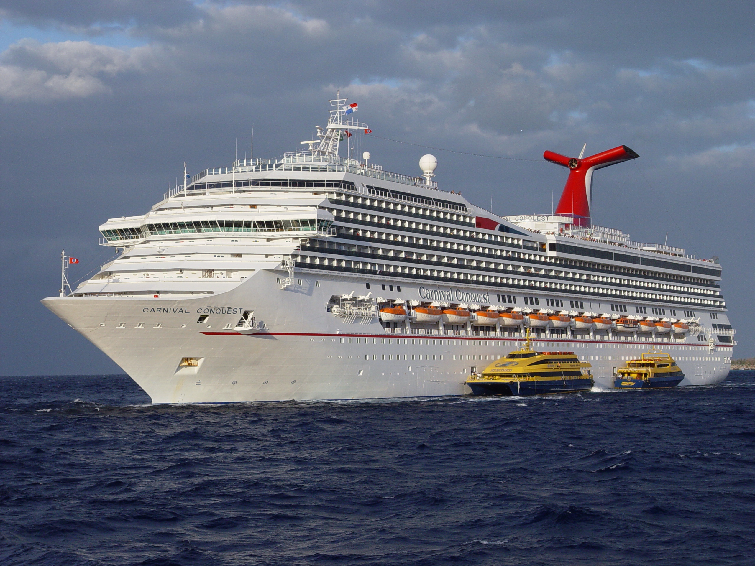 Ein großes weißes Kreuzfahrtschiff mit dem Namen Carnival Conquest fährt auf dem Meer, begleitet von zwei gelben Booten.