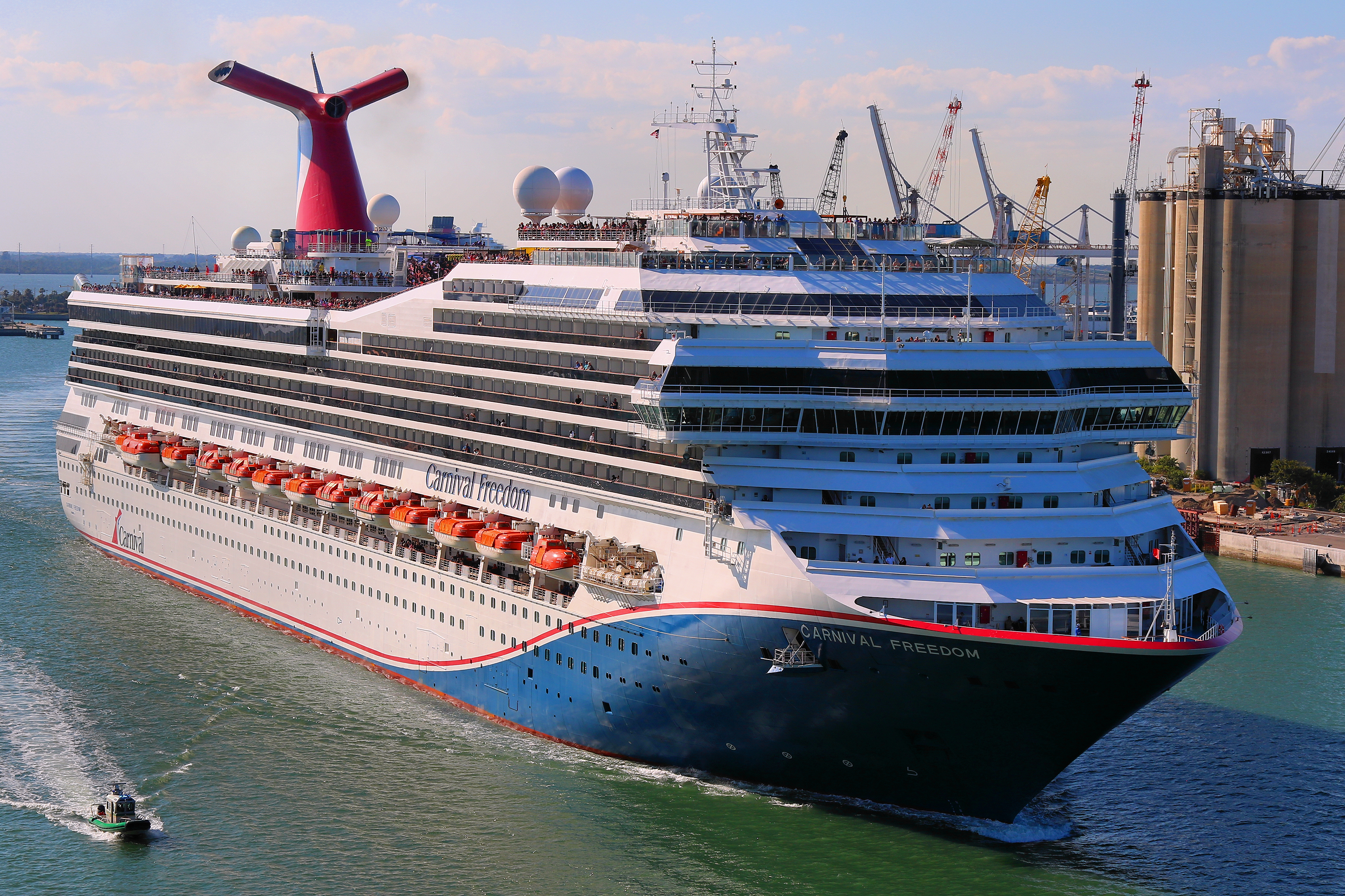 Ein großes Kreuzfahrtschiff mit dem Namen Carnival Freedom fährt auf dem Wasser, im Hintergrund sind Kräne und Silos zu sehen.