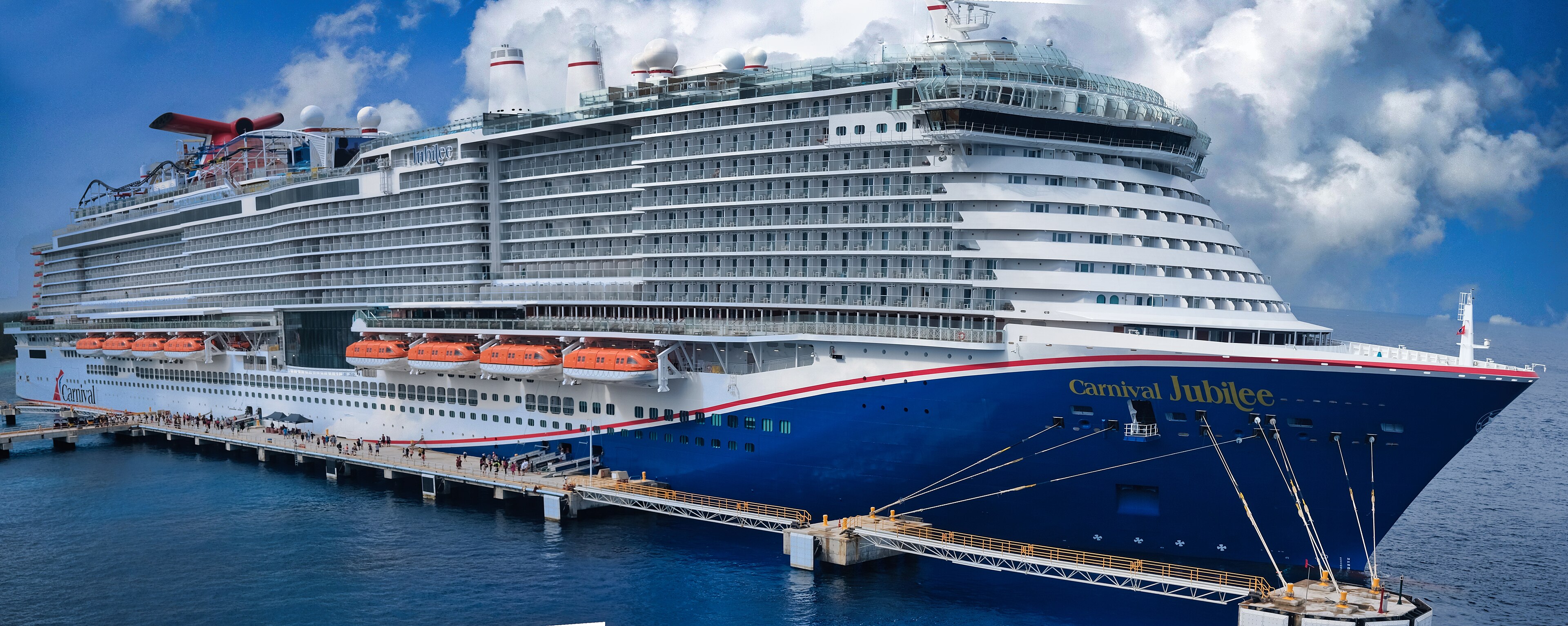 Ein großes blau-weißes Kreuzfahrtschiff namens Carnival Jubilee liegt an einem Pier im Meer, Menschen gehen über den Gang an Bord.