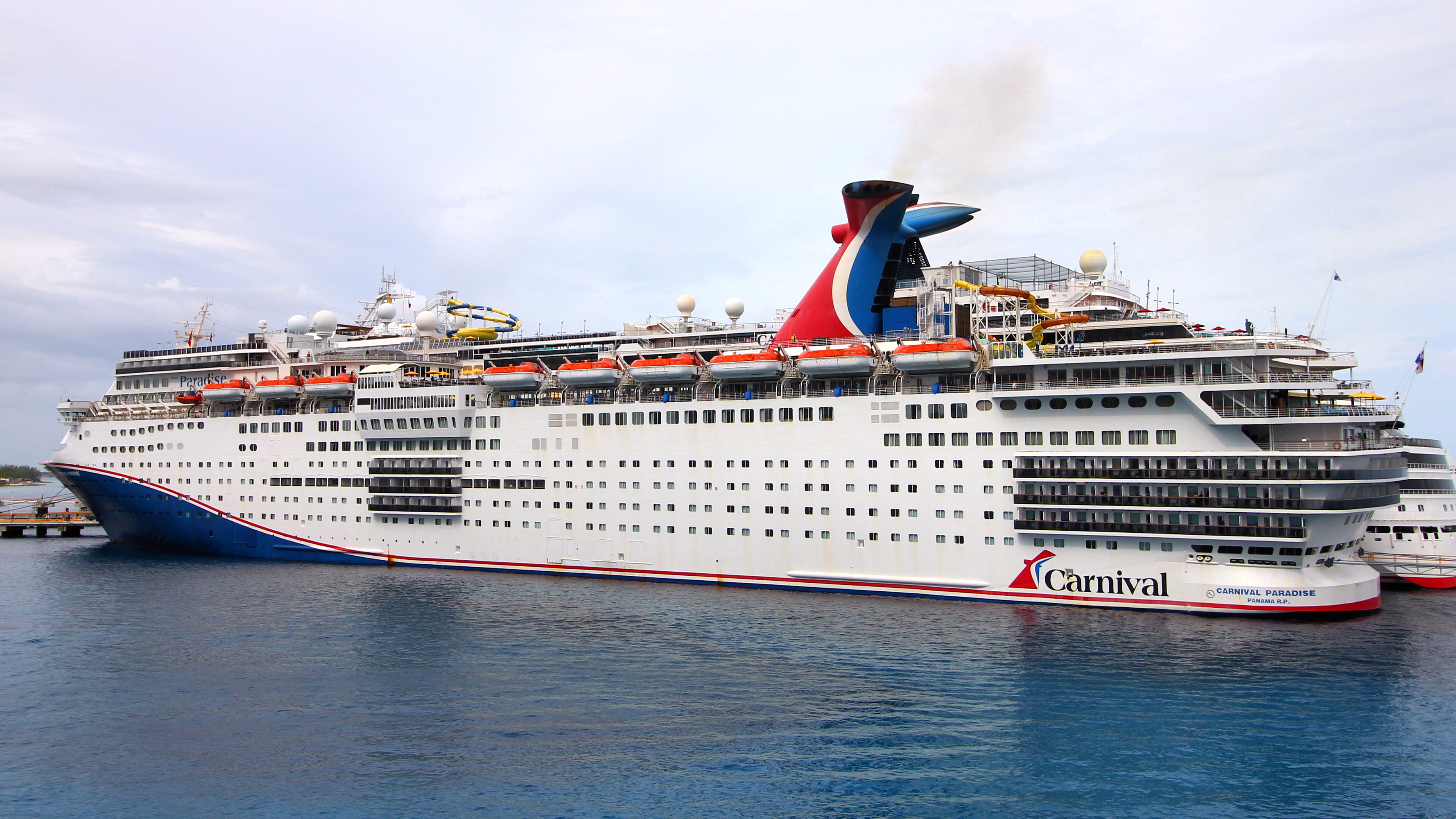 Ein großes weißes Kreuzfahrtschiff mit dem Namen Carnival Paradise liegt im Wasser vor Anker.