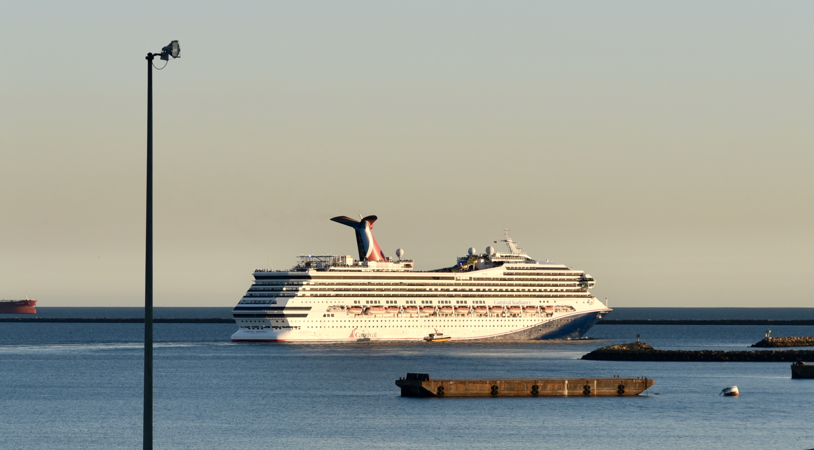 Ein großes weißes Kreuzfahrtschiff mit dem Namen Carnival Radiance fährt in einen Hafen ein, im Vordergrund ist ein Ponton zu sehen.