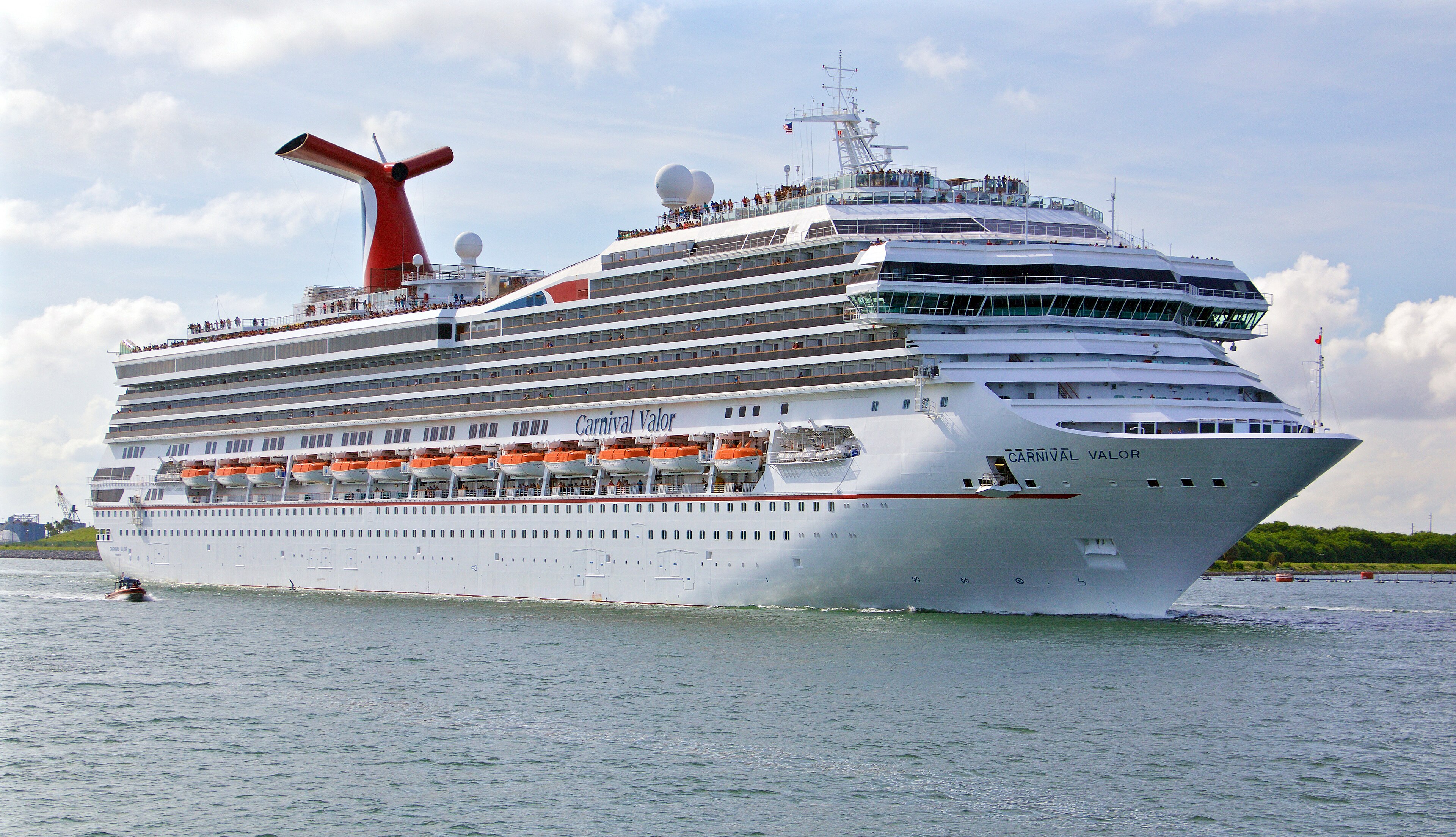 Ein großes weißes Kreuzfahrtschiff namens Carnival Valor fährt auf dem Wasser, mit vielen Passagieren auf den Decks und Rettungsbooten an der Seite.