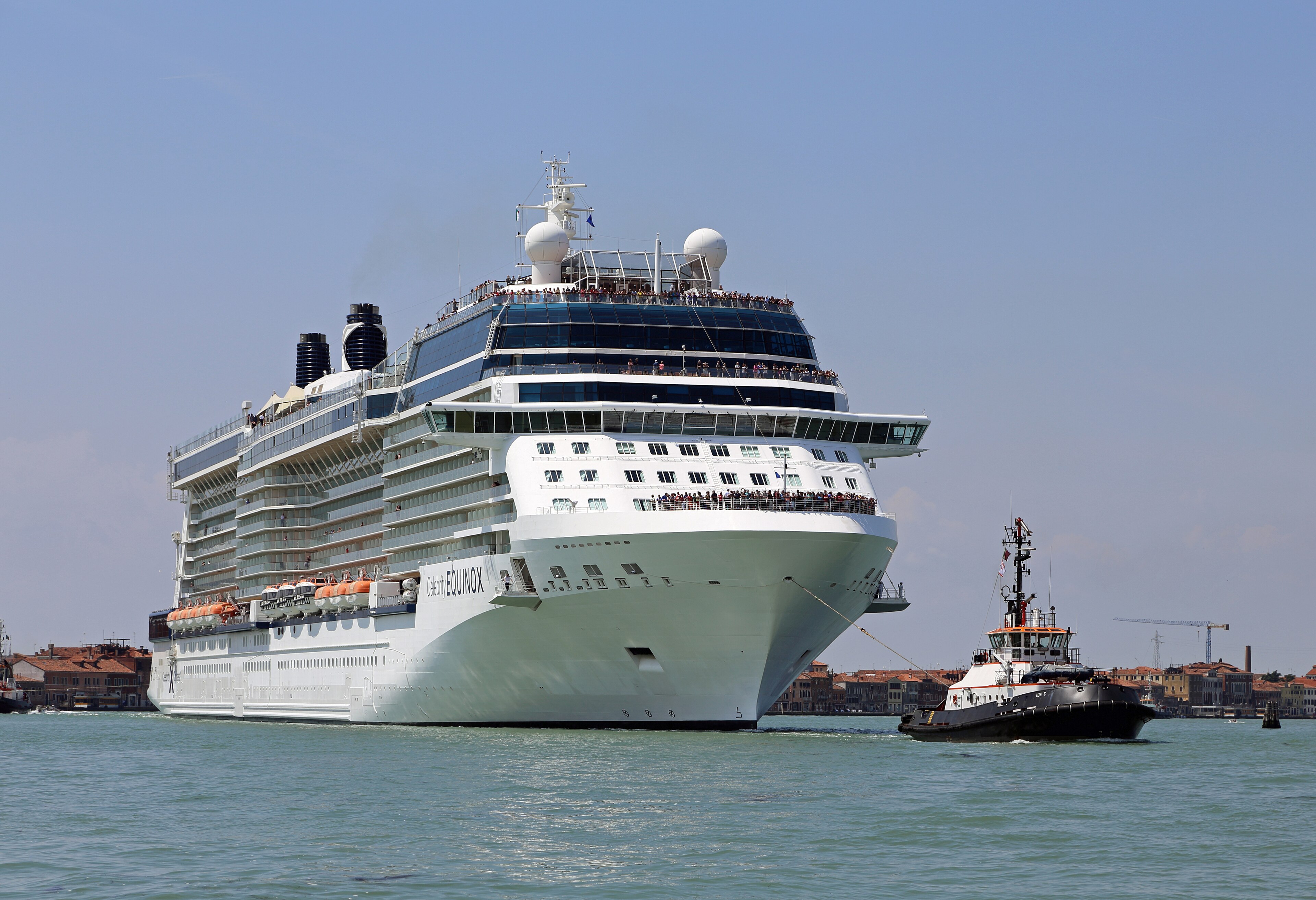 Ein großes Kreuzfahrtschiff namens Celebrity Equinox wird von einem Schlepper durch das Wasser gezogen, im Hintergrund ist eine Stadt mit Gebäuden zu sehen.