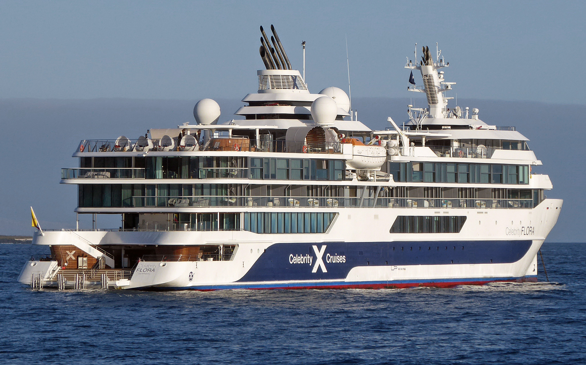 Ein grosses weisses Kreuzfahrtschiff mit dem Namen Celebrity Flora faehrt auf dem blauen Meer.