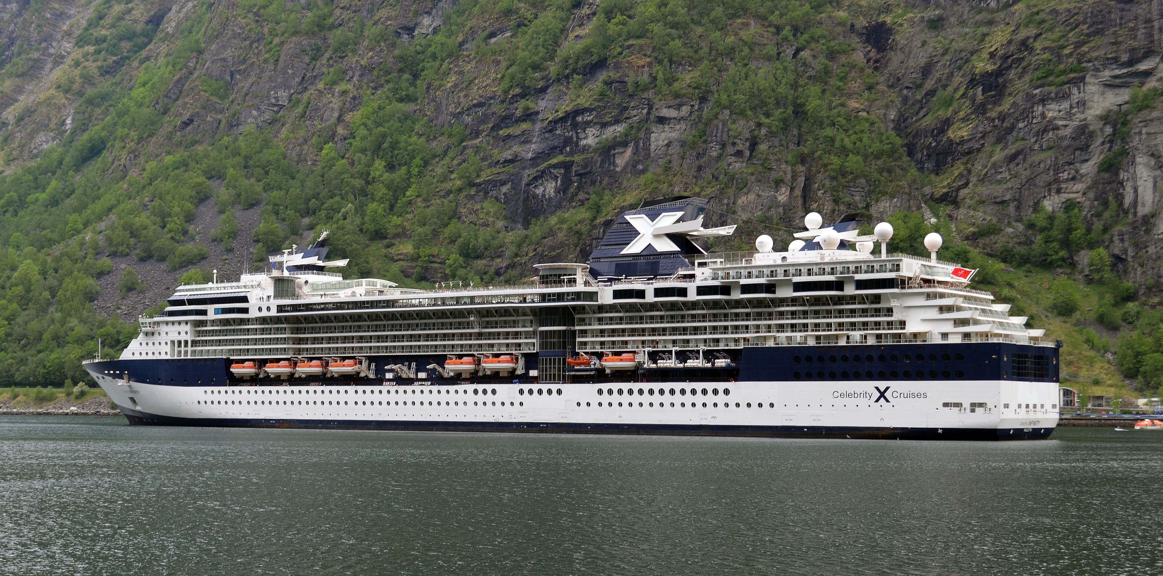 Ein großes Kreuzfahrtschiff der Marke Celebrity Cruises liegt im Wasser vor einer steilen, bewaldeten Felswand.