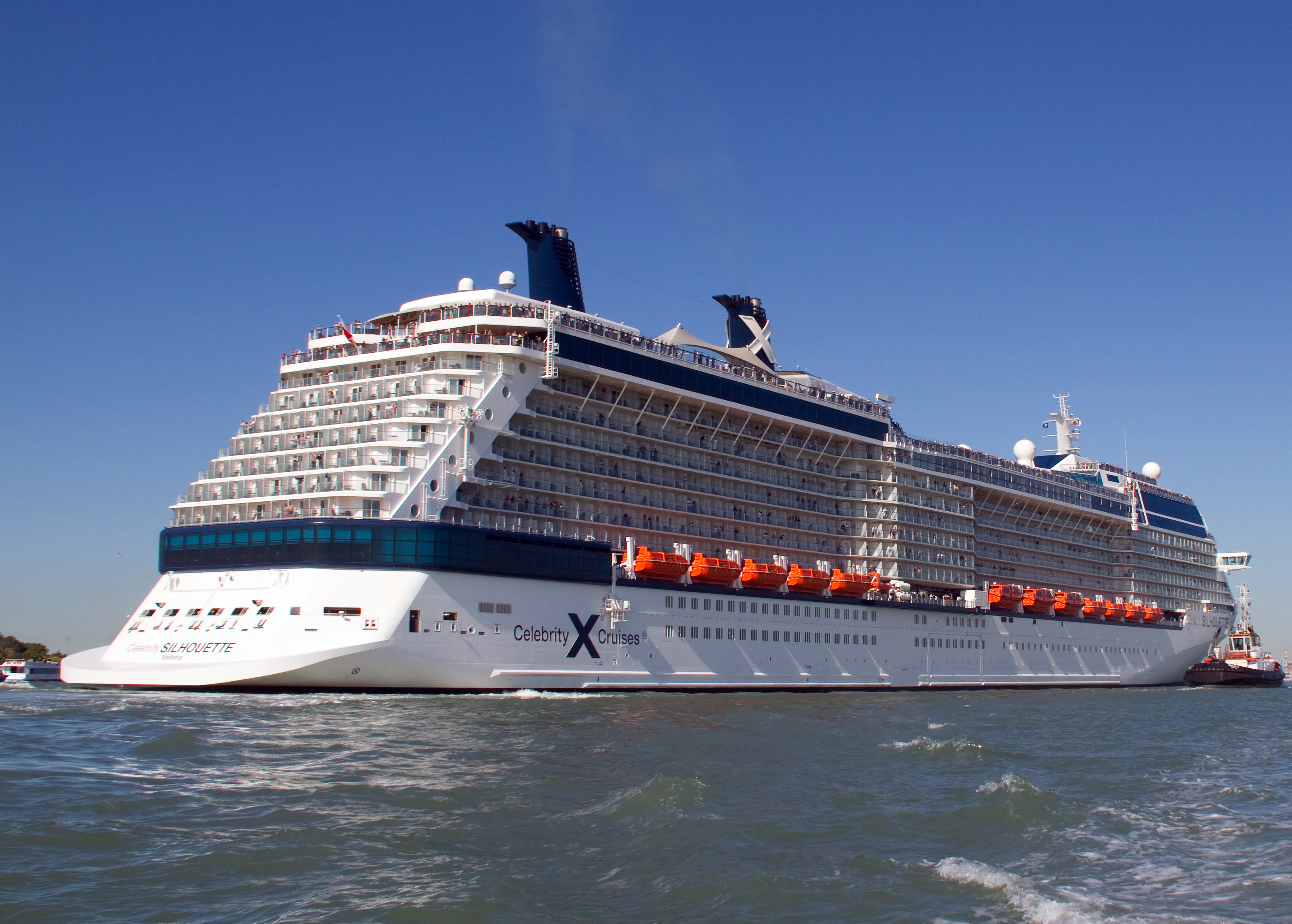 Ein grosses weisses Kreuzfahrtschiff mit dem Namen Celebrity Silhouette faehrt auf dem Wasser, begleitet von einem Schlepper.