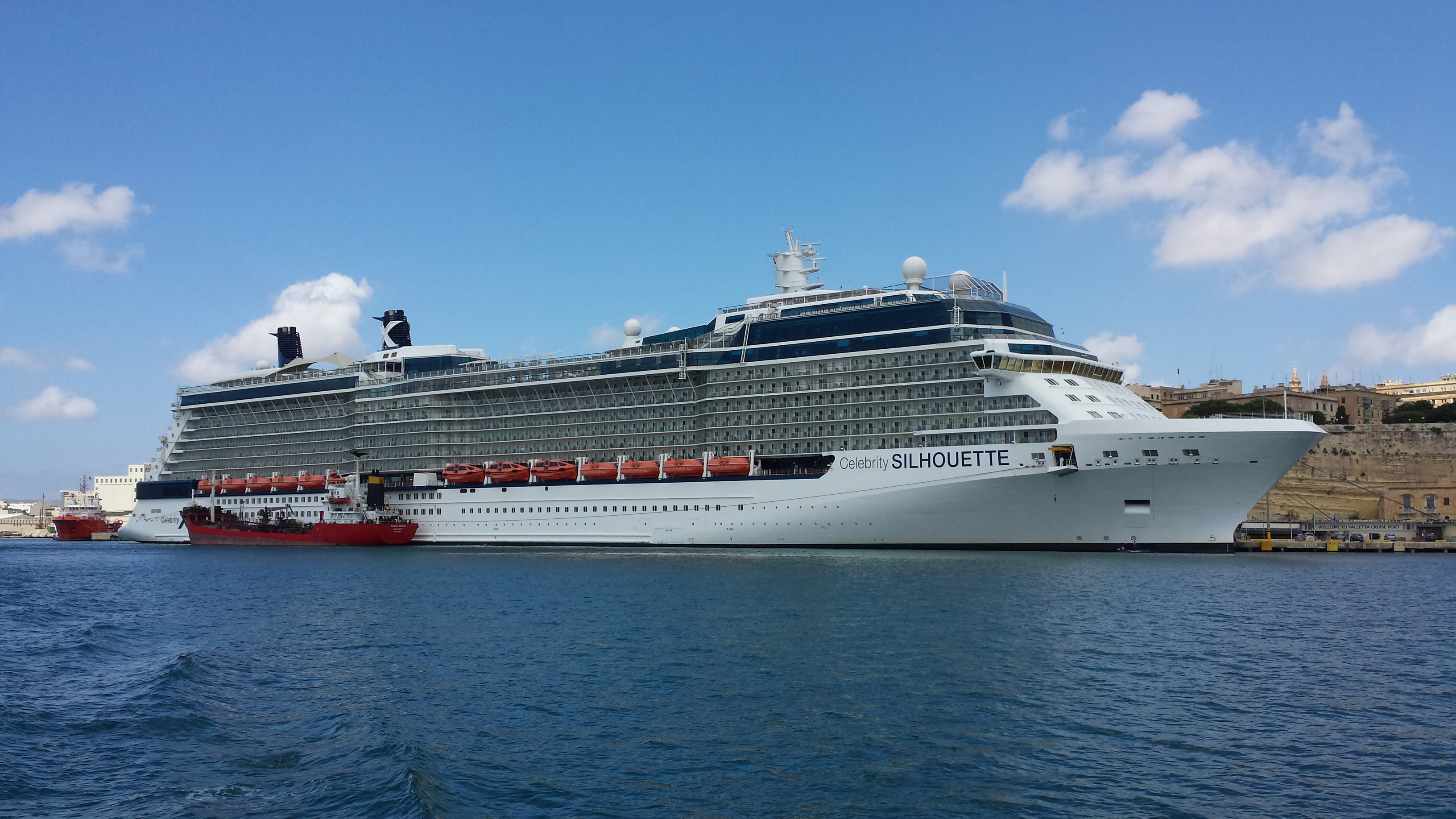 Ein großes Kreuzfahrtschiff namens Celebrity Silhouette liegt im Hafen, daneben ein kleineres rotes Schiff.
