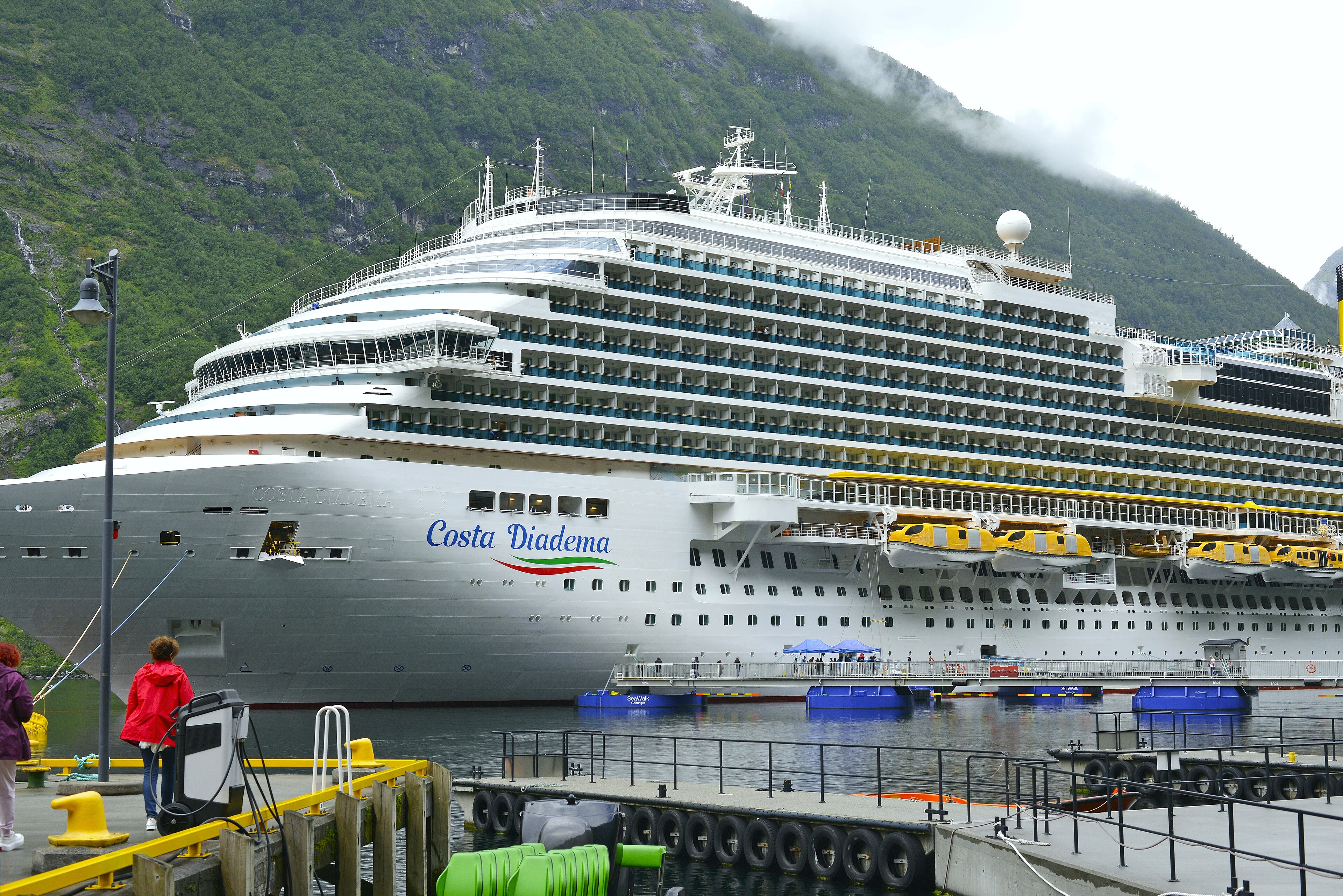 Ein großes Kreuzfahrtschiff namens Costa Diadema liegt im Hafen vor einer bewaldeten Berglandschaft.