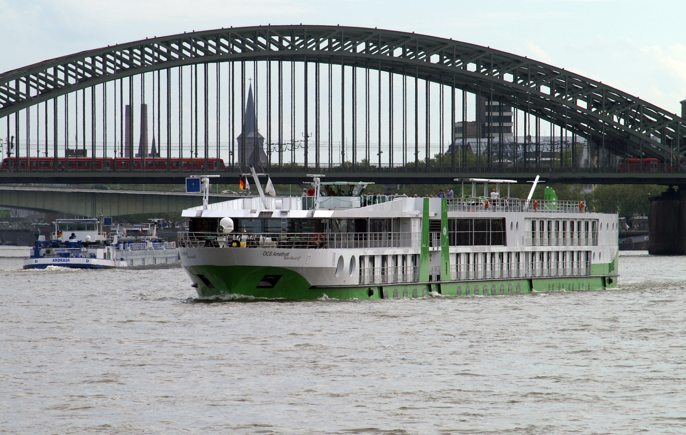 Ein grünes und weißes Flussschiff fährt auf einem Fluss unter einer großen Eisenbahnbrücke hindurch, auf der ein Zug fährt.