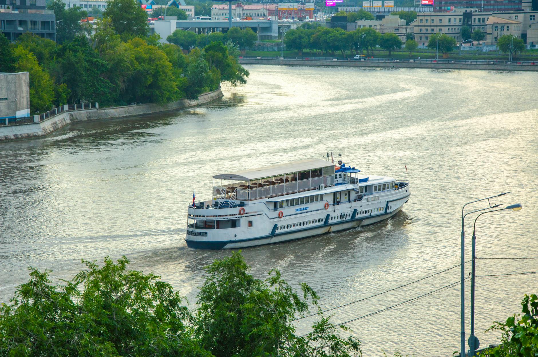 Ein weisses Ausflugsboot faehrt auf einem breiten Fluss, im Hintergrund eine Stadt mit Gruenflaechen.