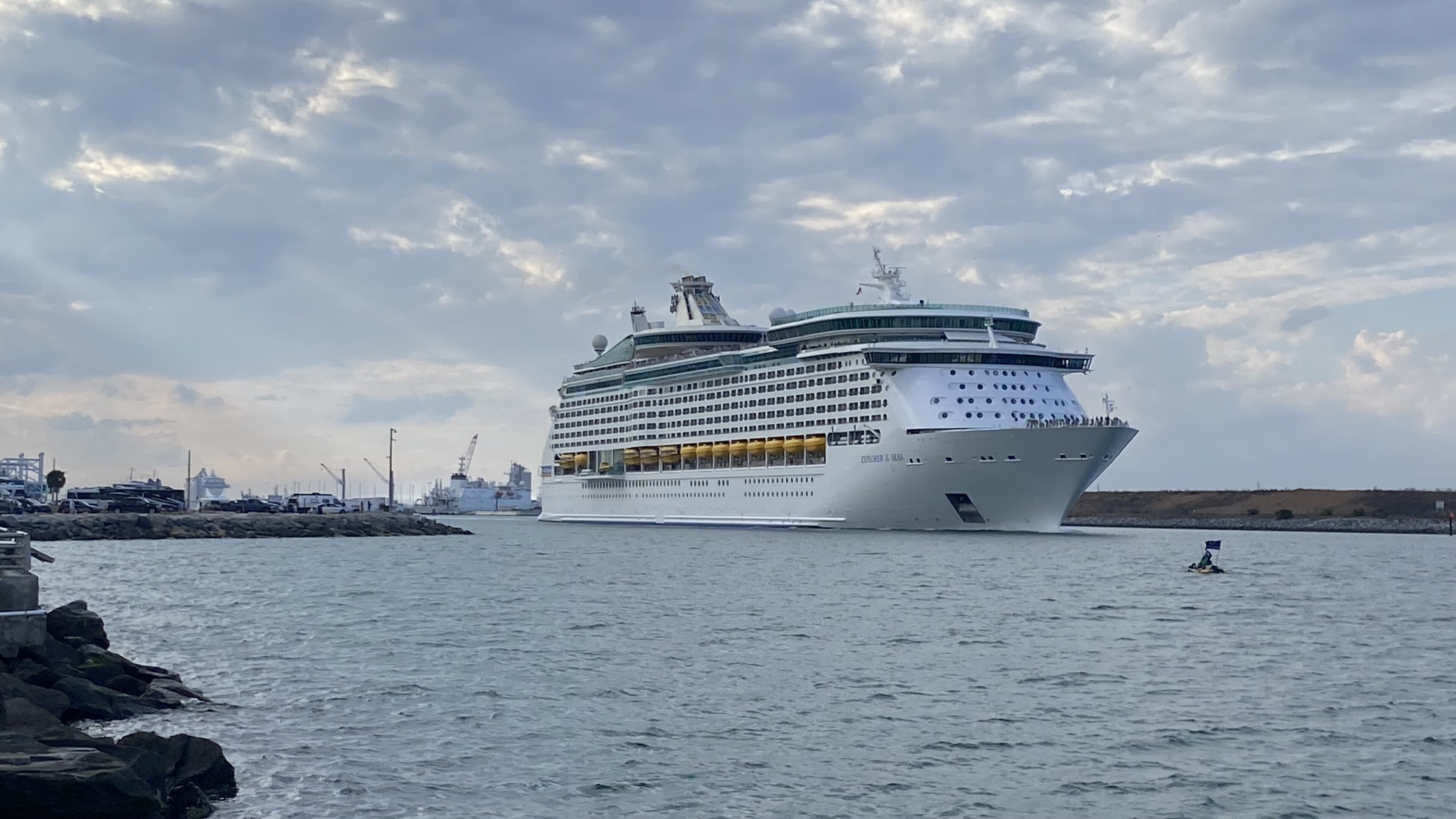 Ein großes weißes Kreuzfahrtschiff fährt auf dem Meer in Richtung des Betrachters, mit einer Hafeneinfahrt im Hintergrund und einer Person auf einem kleinen Boot mit Flagge im Vordergrund.